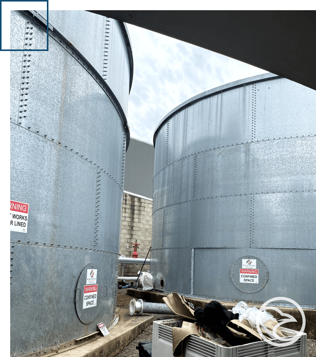 Large industrial storage tanks with warning signs are visible under a clear sky, with various equipment scattered in the foreground.